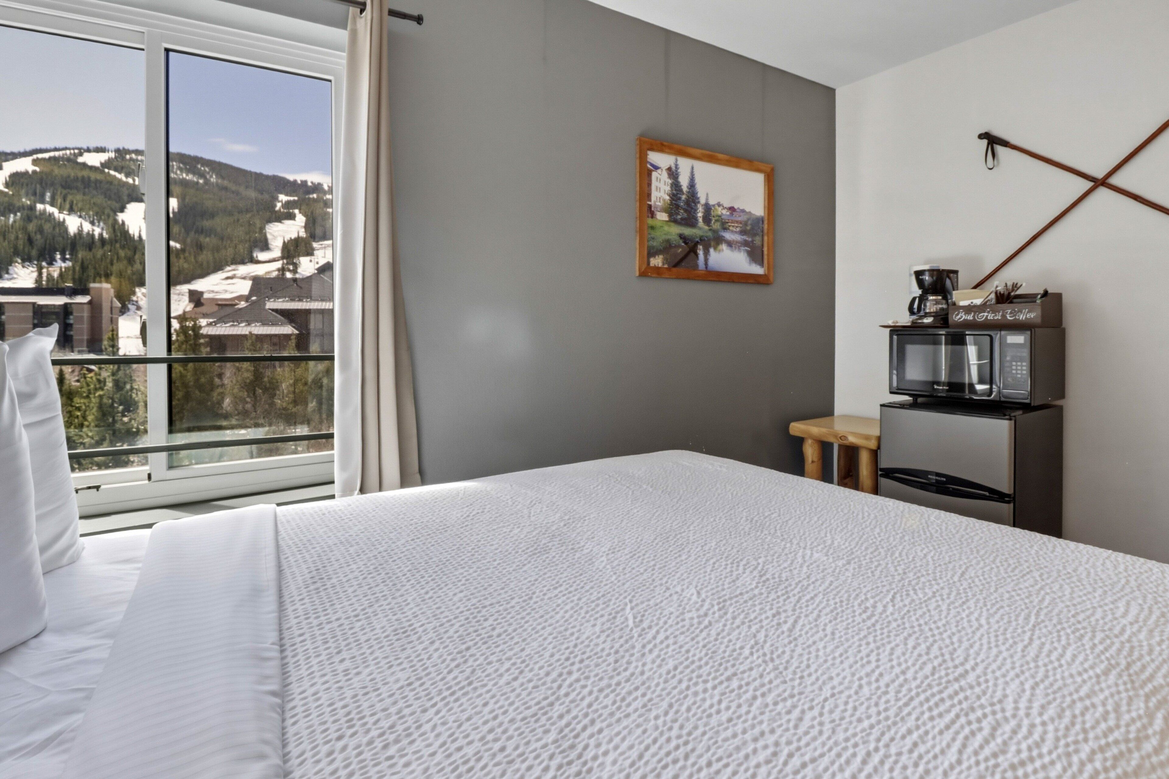 Habitación de hotel con cama, ventana con vista a la montaña, pequeña mesa con cafetera y un cuadro en la pared.