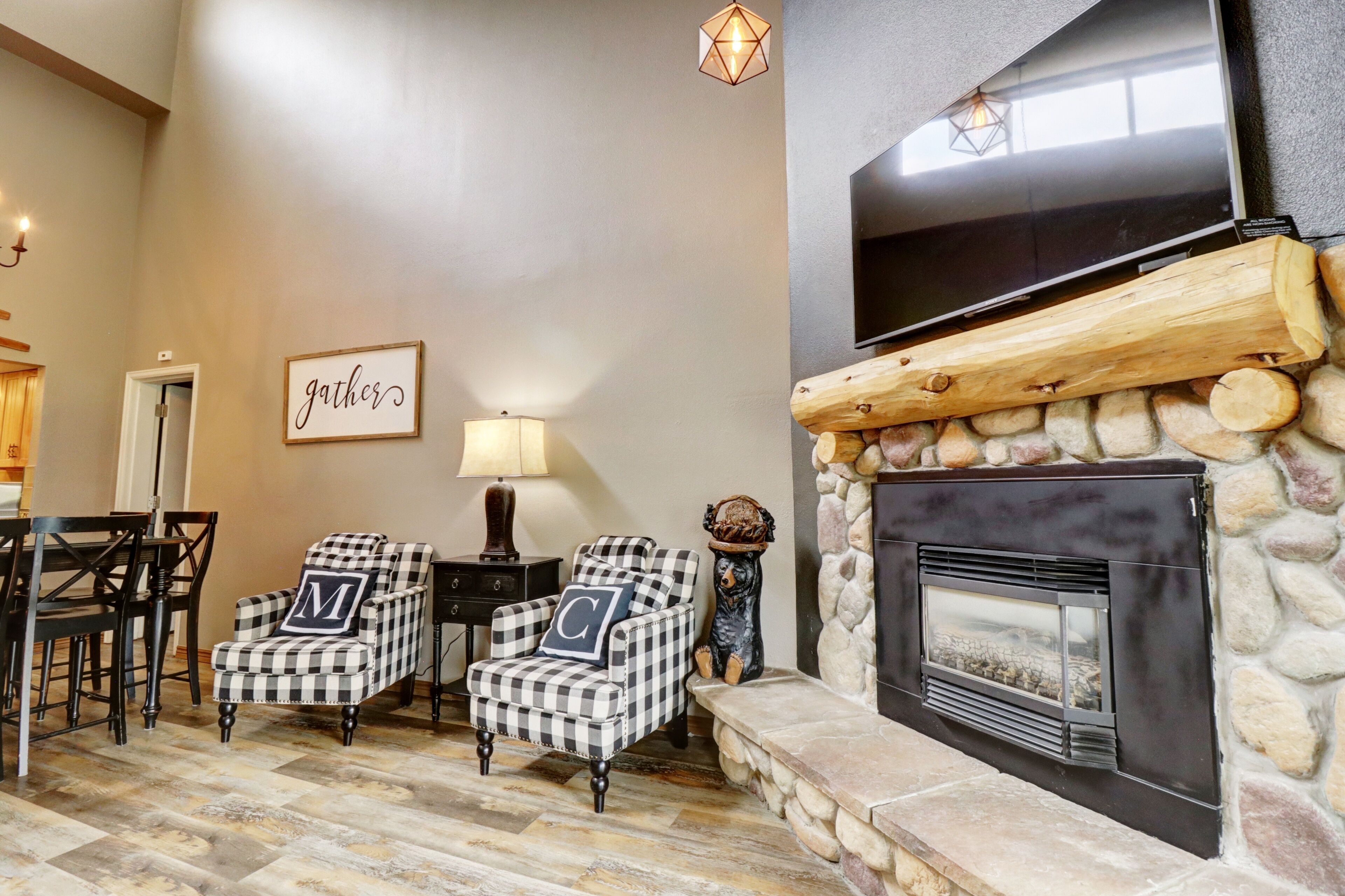 A cozy living room with a stone fireplace, checkered armchairs, a television, and a 'gather' sign on the wall.