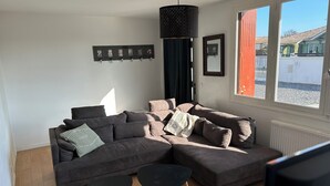 A modern living room with a sectional sofa, two small black tables, a black pendant light, a wall-mounted picture frame, and a window with a view of the outside.