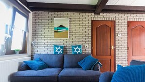 A living room with a grey sofa, two blue pillows, a wooden door, and a brick wall with a painting. - Bungalow in North Holland Coast (Sint Maartensvlotbrug)