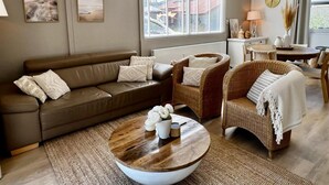 A living room with a brown leather sofa, wicker chairs, a wooden coffee table, and a large window with blinds. - Vacation Bungalow for 4 Persons (Sint Maartensvlotbrug)