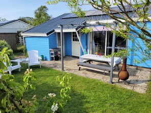 A blue house with a covered patio, a white chair, a table, and a large vase. - Vacation Bungalow for 4 Persons (Sint Maartensvlotbrug)