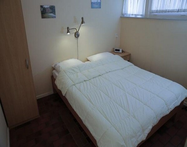 A single bed with a white duvet in a room with a wooden floor, a bedside table with a clock, and a wall-mounted lamp. - Beautiful Bungalow in North Holland (Sint Maartensvlotbrug)