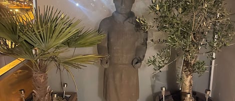 A statue of a figure in a traditional outfit, a potted palm tree, and a large potted tree with metal supports.