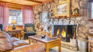 A cozy living room with a stone fireplace, wooden furniture, and a checkered window treatment.