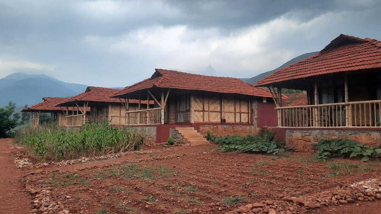 Un edificio rústico de madera con techo de tejas rojas, rodeado por un sendero de tierra y vegetación, con montañas al fondo.