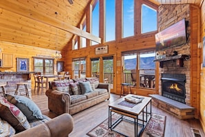 A spacious lodge with a large living area featuring a fireplace, wooden walls, and large windows offering mountain views. - Awesome Endeavor (Sevierville)