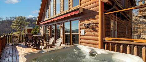 A wooden cabin with a hot tub on the deck, large windows, and a dining area with chairs.