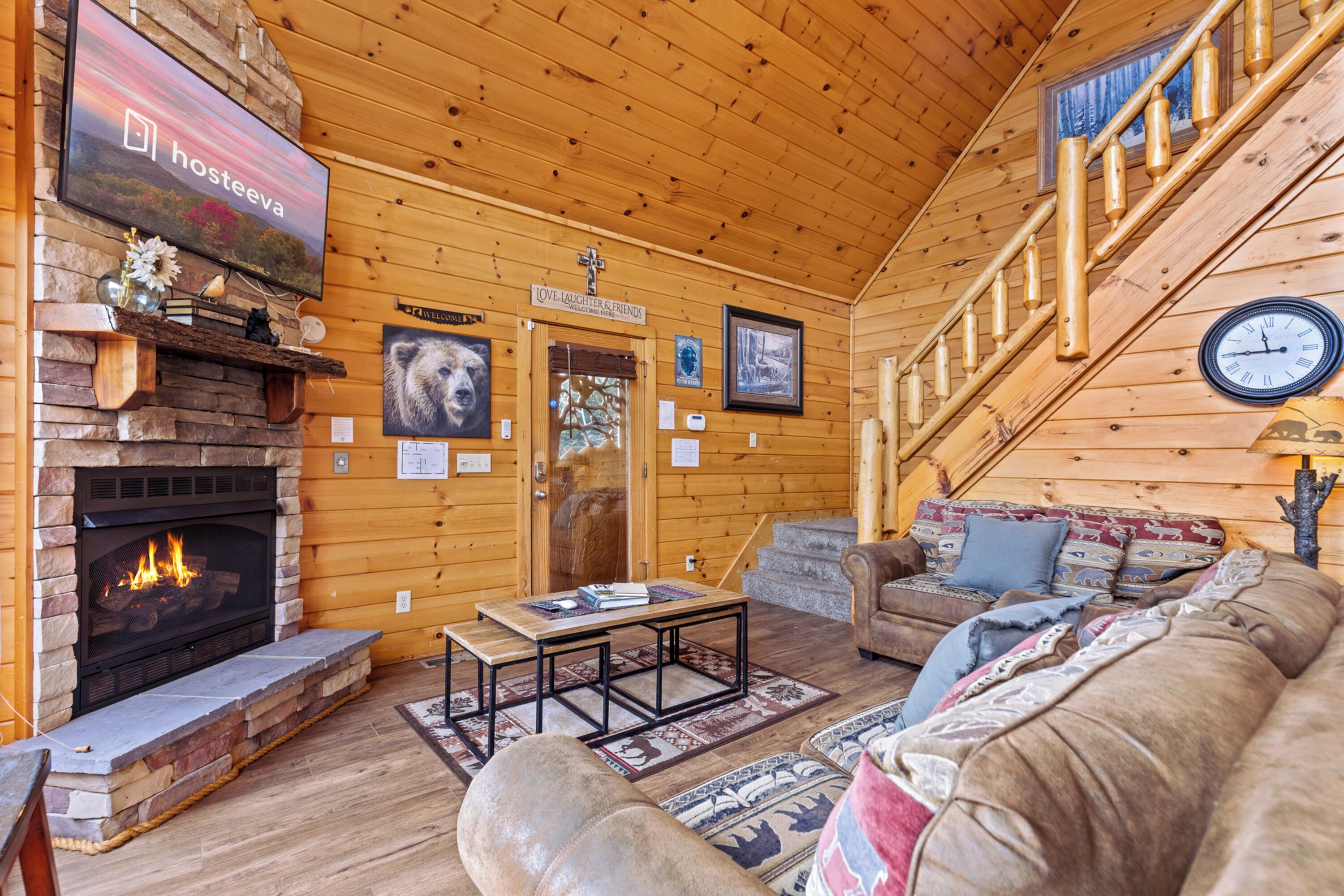 A cozy log cabin interior with a fireplace, a couch, a staircase, and a clock on the wall.