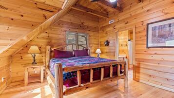 Un dormitorio de cabaña de madera con techo inclinado, una cama con estructura de madera, dos mesitas de noche con lámparas y una ventana con contraventanas.