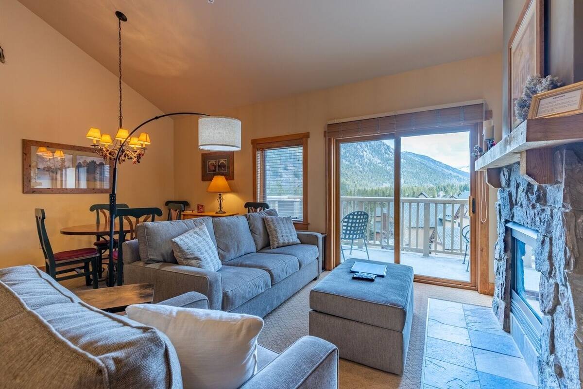 A cozy living room with a grey sofa, a coffee table, and a dining area with a table and chairs. A large window offers a view of mountains.