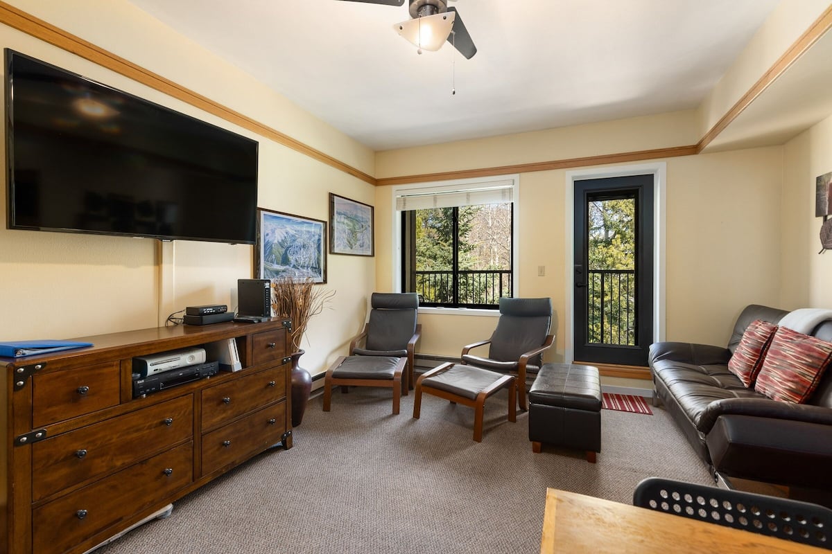 A living room with a flat-screen TV, two armchairs, a sofa, a wooden cabinet, and a window with a view of trees.
