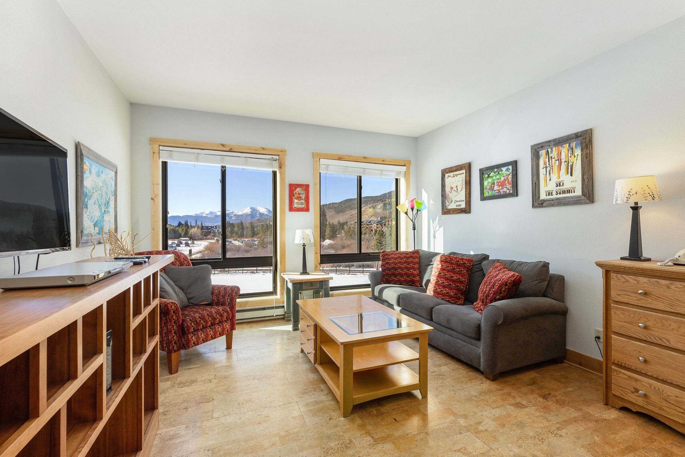 A modern living room with a wooden coffee table, a gray sofa, and a TV on a wooden stand. There are two windows with mountain views, and a wooden cabinet with drawers.