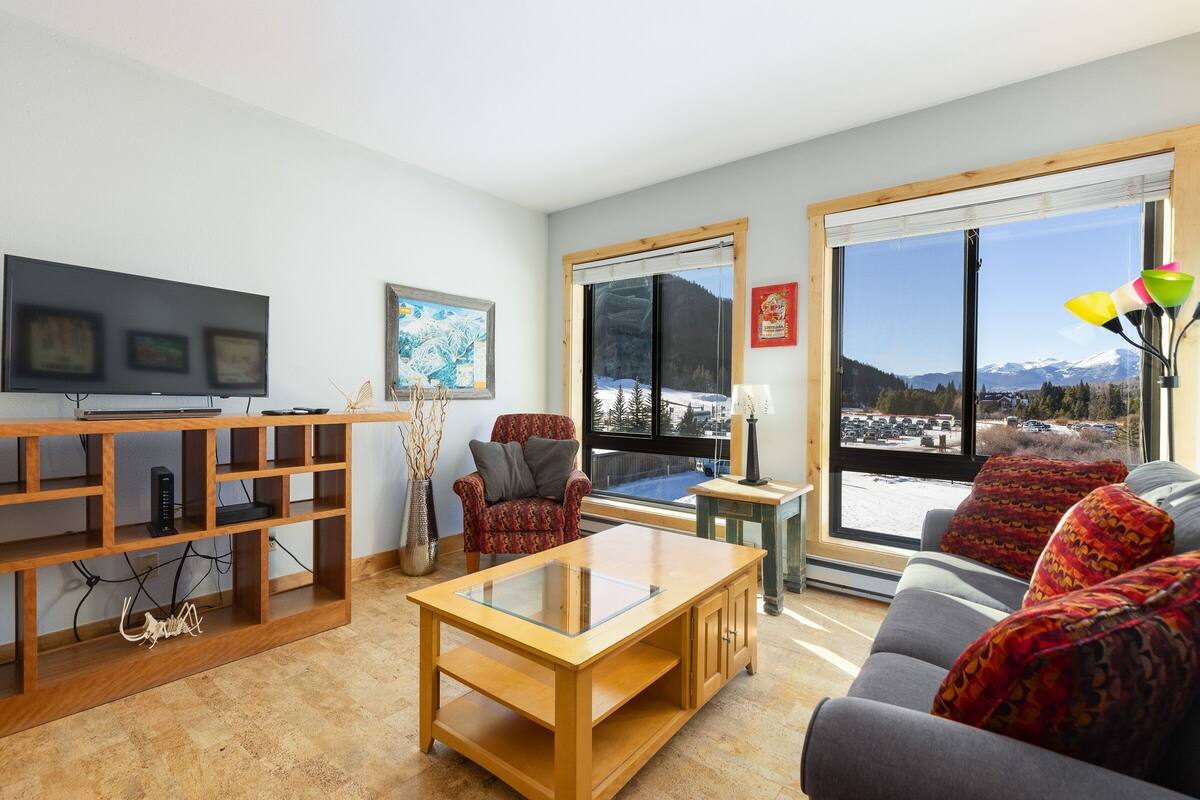 A modern living room with a wooden TV stand, a glass-top coffee table, a sofa with patterned cushions, and a large window offering a mountain view.