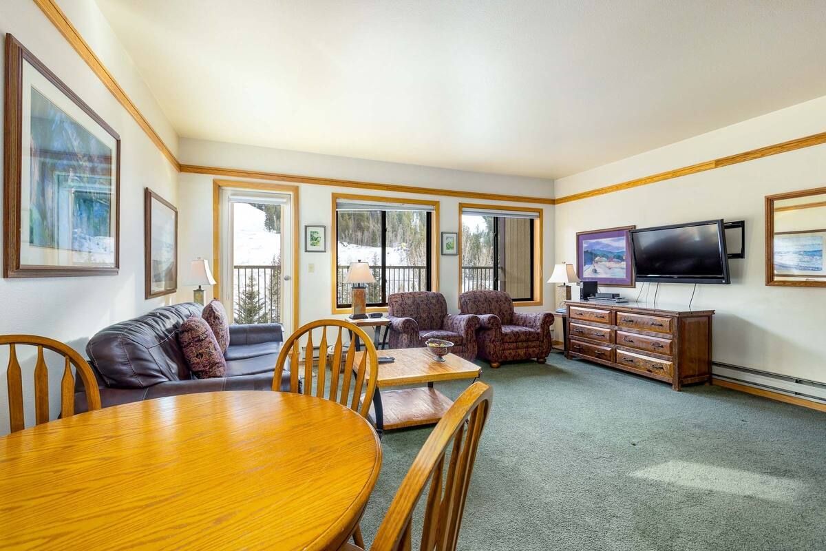 A living room with a wooden dining table, leather sofa, patterned armchairs, a TV, and a wooden cabinet.