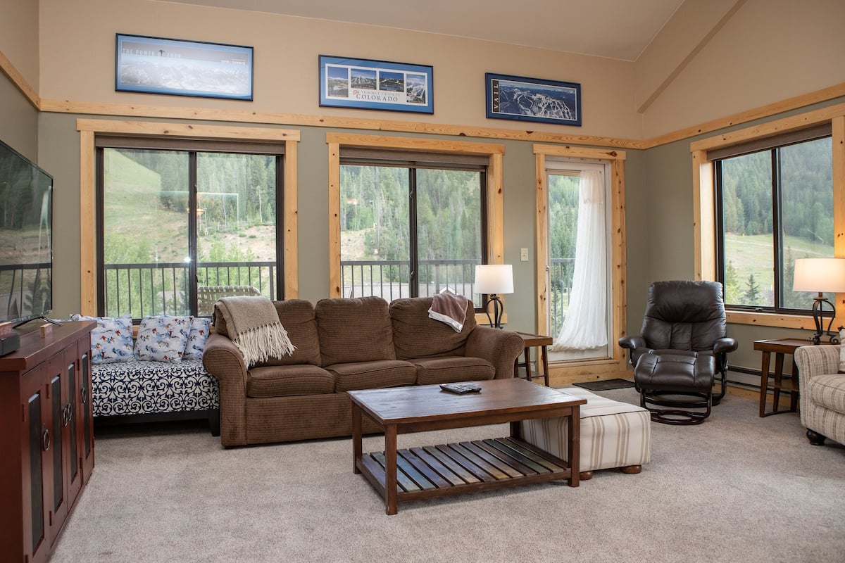 A cozy living room with a brown sofa, a coffee table, a recliner, and a TV on a wooden cabinet. Large windows offer views of a forested area.