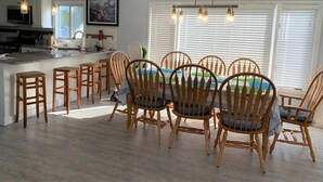 Un coin repas avec des chaises en bois et une table centrale, une cuisine avec des meubles blancs et une grande fenêtre équipée de stores.