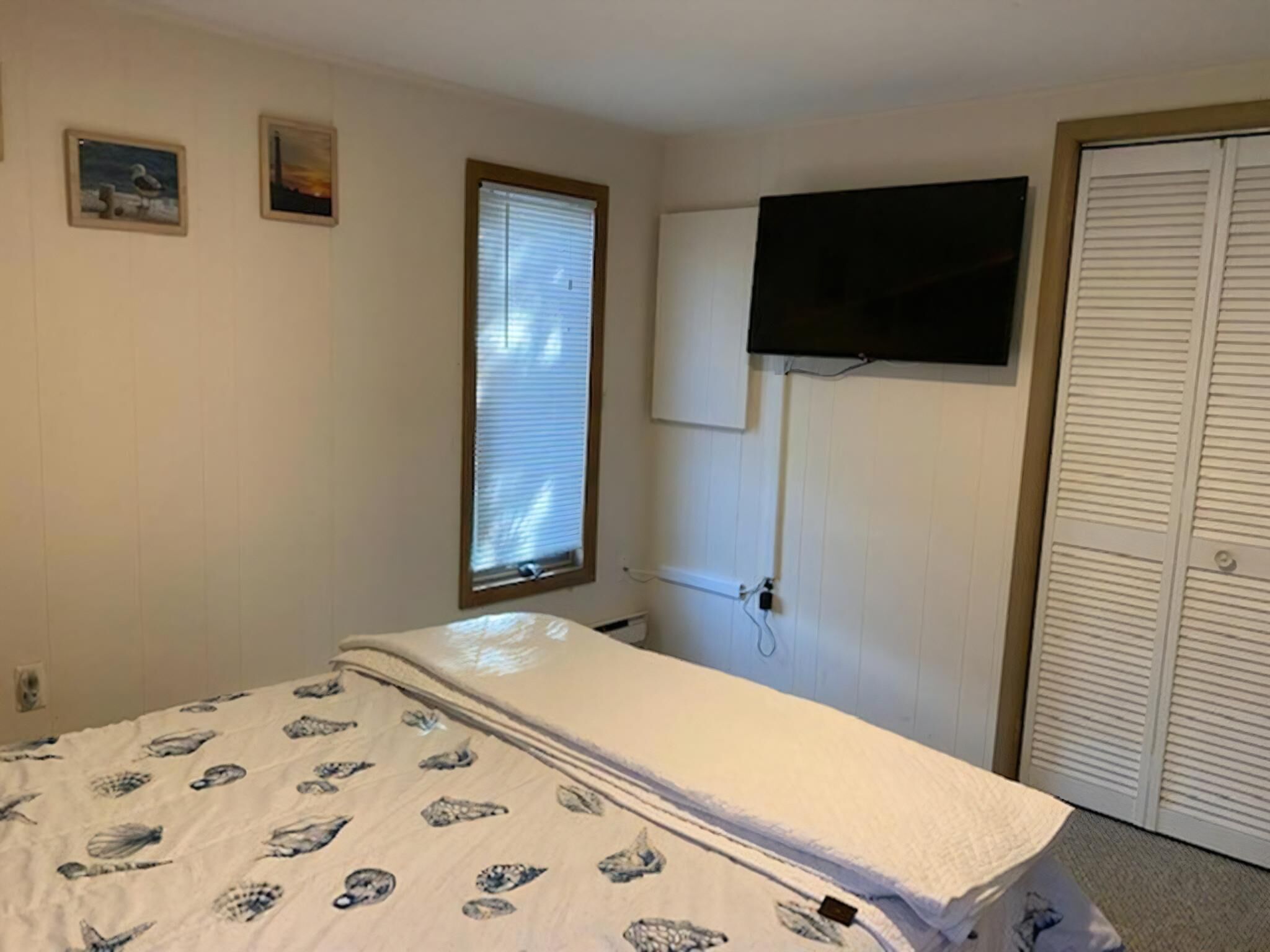 A bedroom with a bed, a window with blinds, a TV mounted on the wall, and a closet.