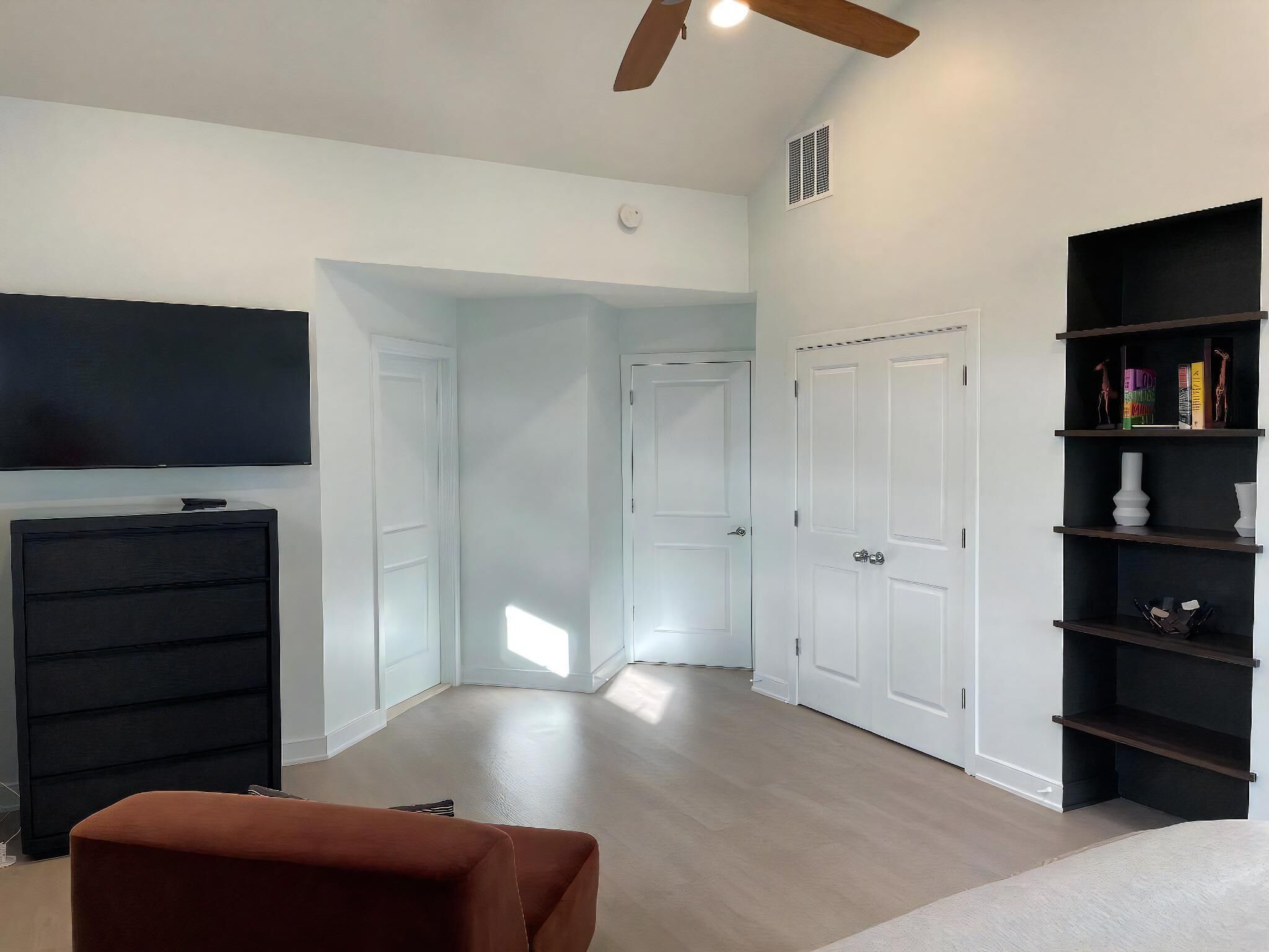 Ein Zimmer mit Deckenventilator, Flachbildfernseher, schwarzem Bücherregal und braunem Pouf.