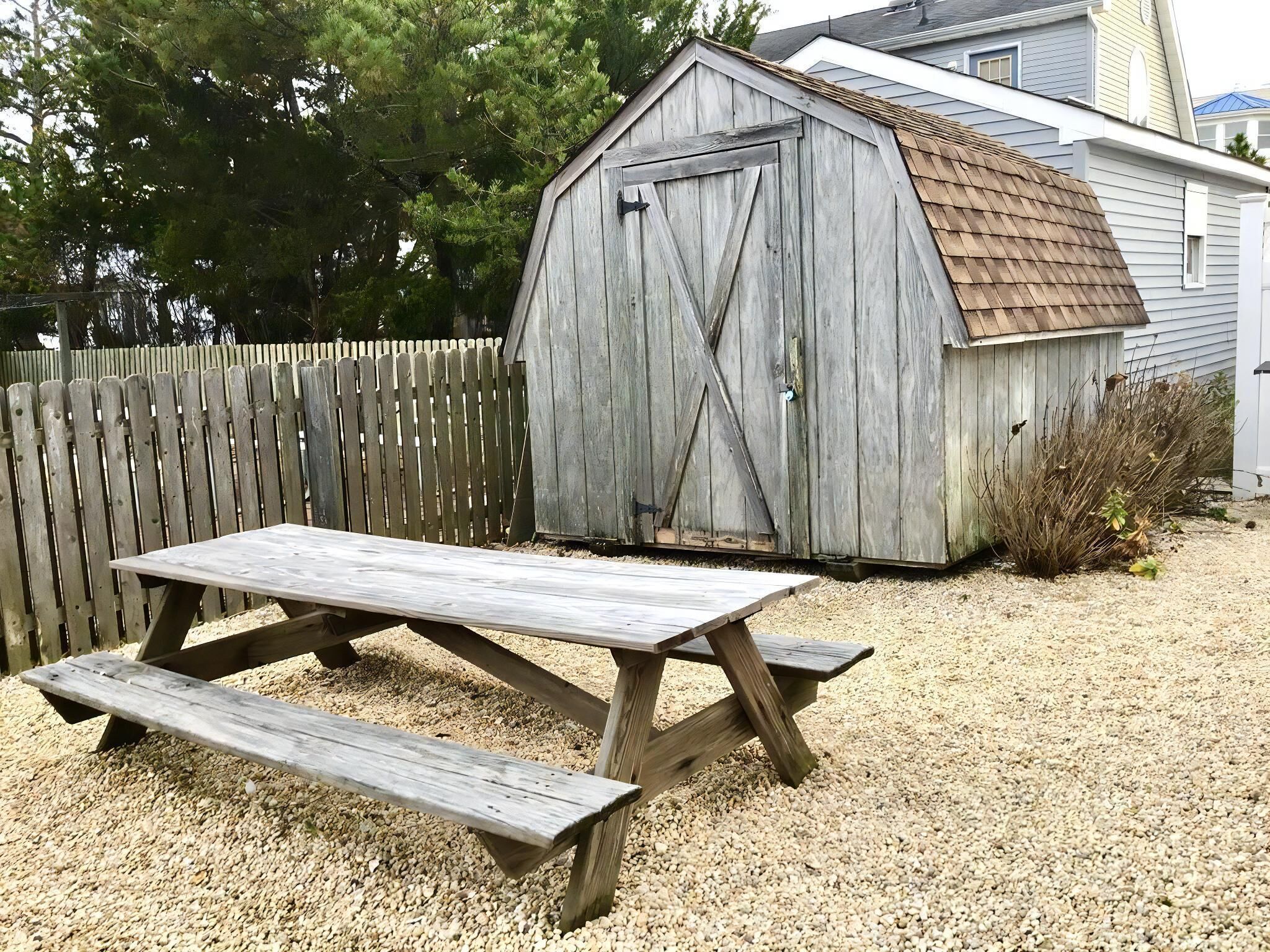 A wooden shed with a sloped roof and a door, a picnic table with benches, and a wooden fence in the background.