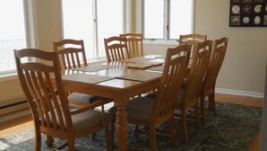 Una mesa de comedor con seis sillas de madera, un tapete estampado y una pared decorada con motivos náuticos enmarcados.