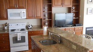 Una cocina con armarios de madera, microondas, estufa, encimera de granito, fregadero y televisor.