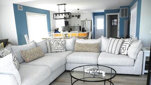 A modern living room with a sectional sofa, a glass coffee table, and a kitchen in the background. - 4 bedroom accommodation in Harvey Cedars (Harvey Cedars)