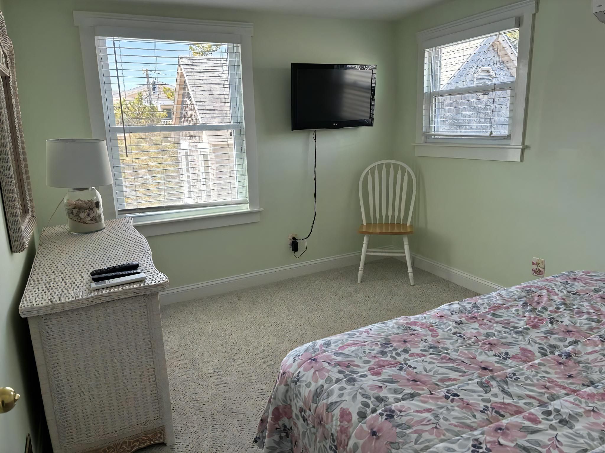 Un dormitorio con cama, una silla, televisor montado en la pared y una ventana con persianas.