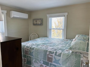 A bedroom with a bed, a chair, a dresser, a window, and an air conditioning unit.