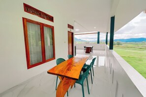A dining area with a wooden table and chairs, large windows with wooden frames, and a view of a green landscape.