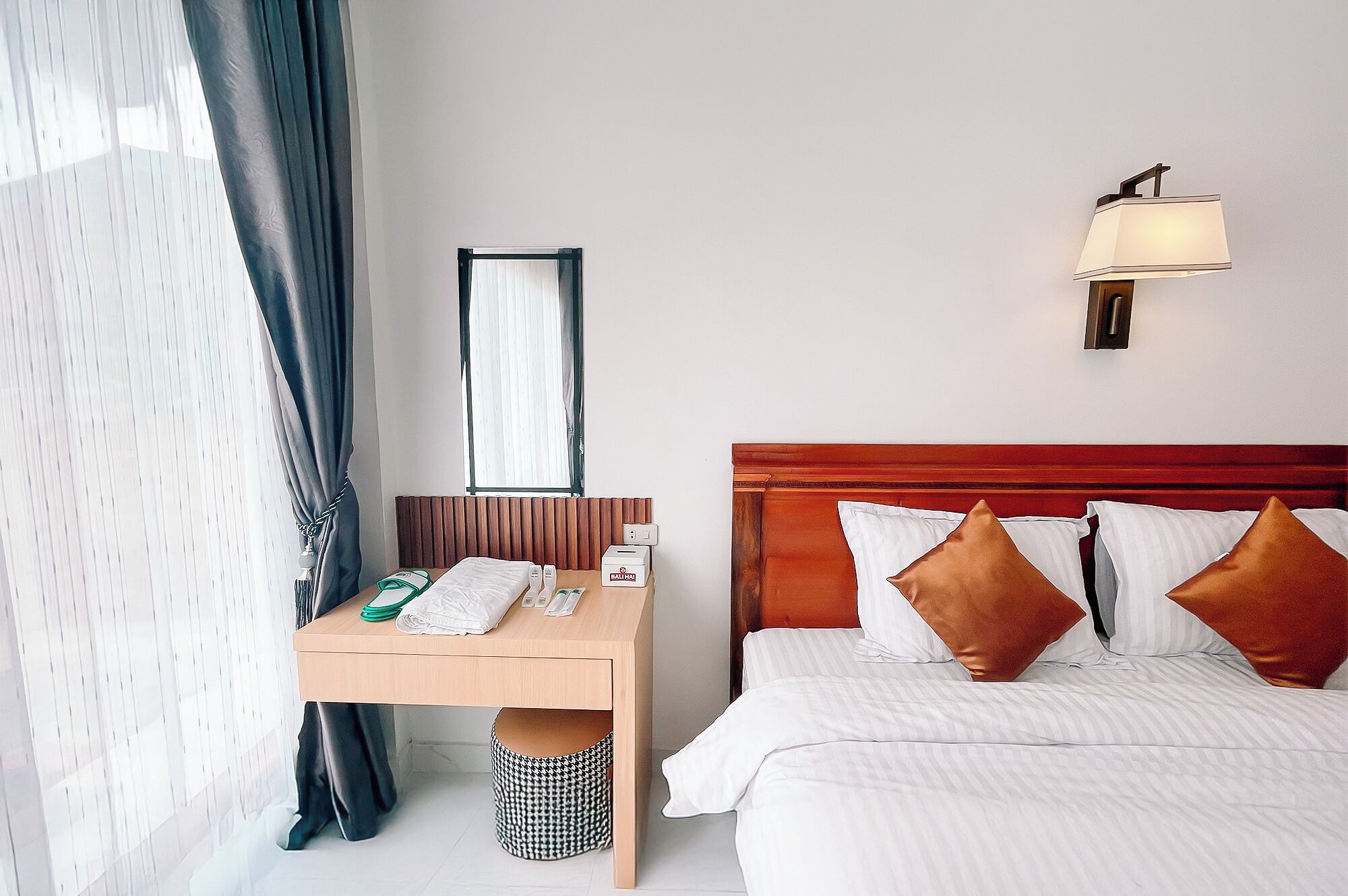 A hotel room with a wooden desk, a bed with white and brown pillows, a nightstand, a lamp, and a window with curtains.