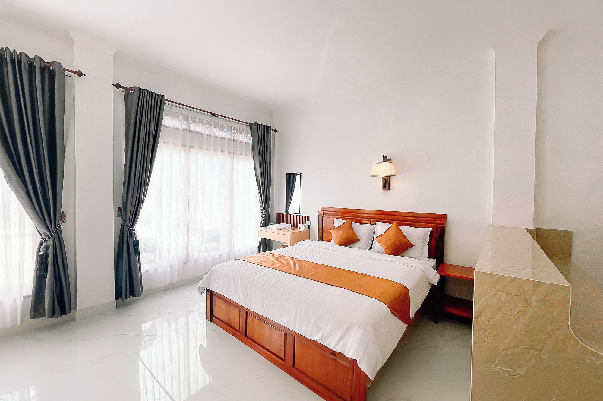 A hotel room with a large bed, a bedside table, a lamp, and a view of the outside through sheer curtains.