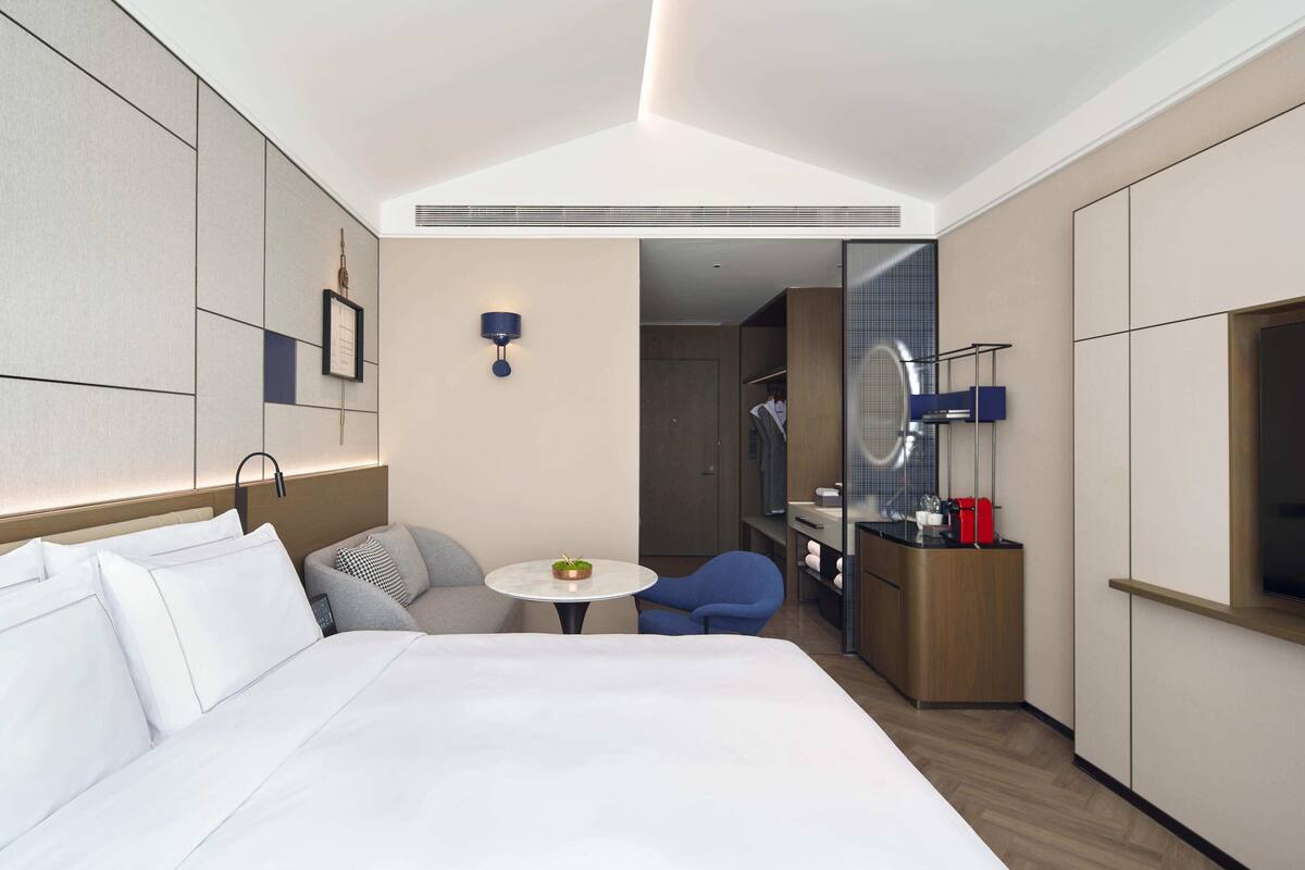 Une chambre d’hôtel avec un grand lit, une petite table ronde ornée d’une plante, une chaise bleue, un minibar et un miroir.