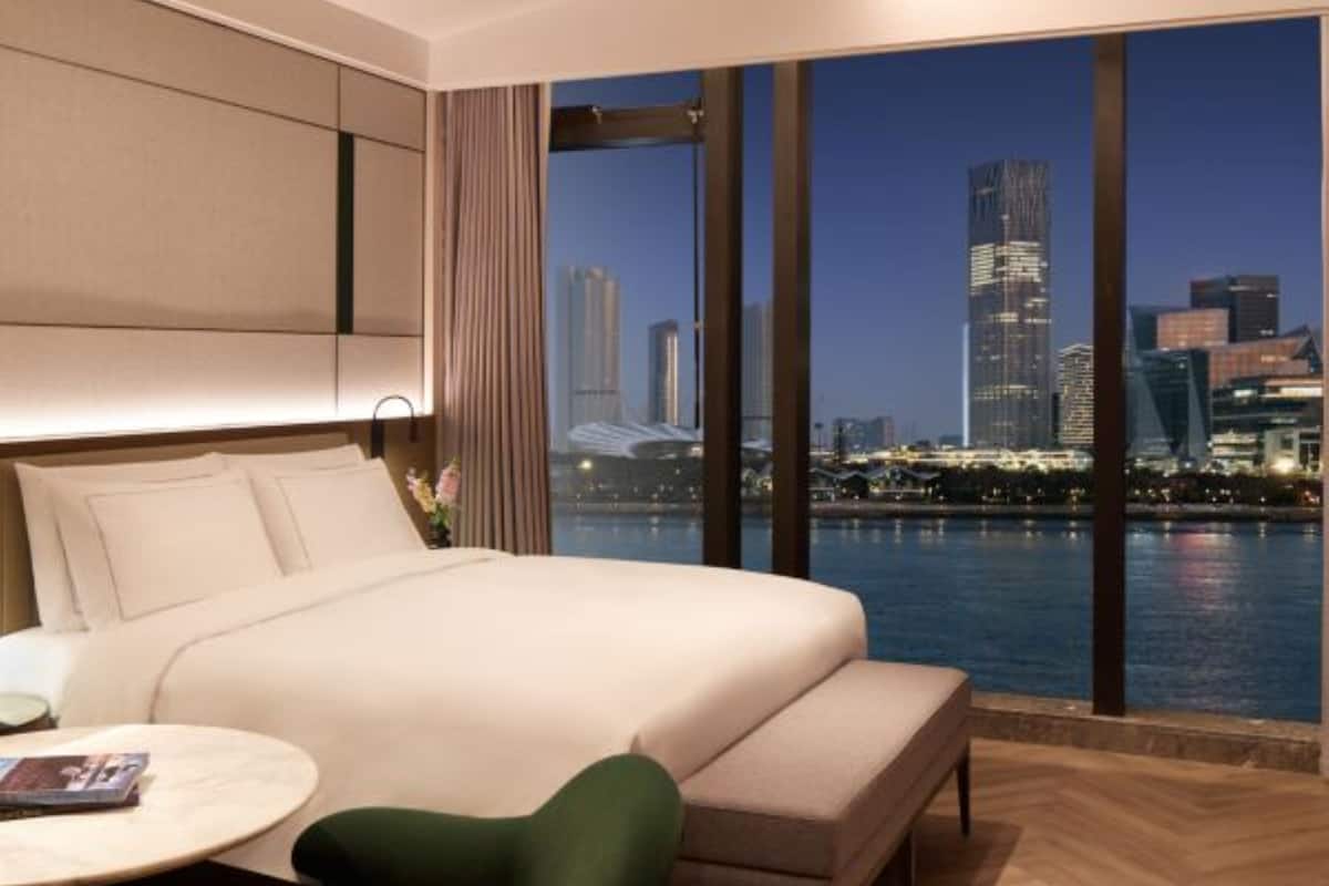 Une chambre d’hôtel moderne avec un grand lit, une petite table et une chaise. De grandes fenêtres offrent une vue sur le paysage urbain.