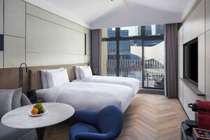 Habitación de hotel con dos camas, una mesita con fruta, una silla azul, un televisor y un balcón con vista a la ciudad.