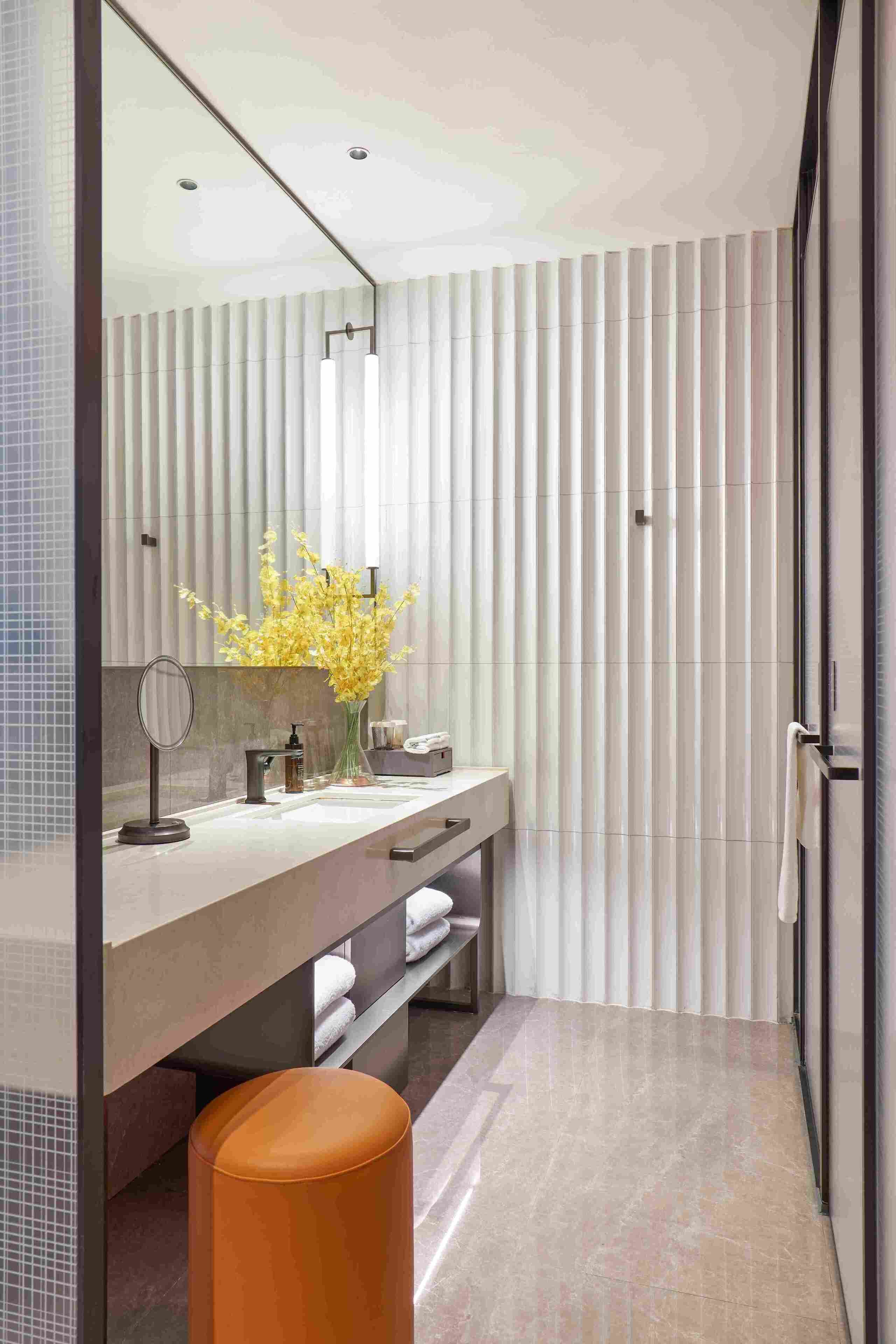 A modern bathroom with a large mirror, a vanity with a sink, a vase of yellow flowers, and a towel rack.