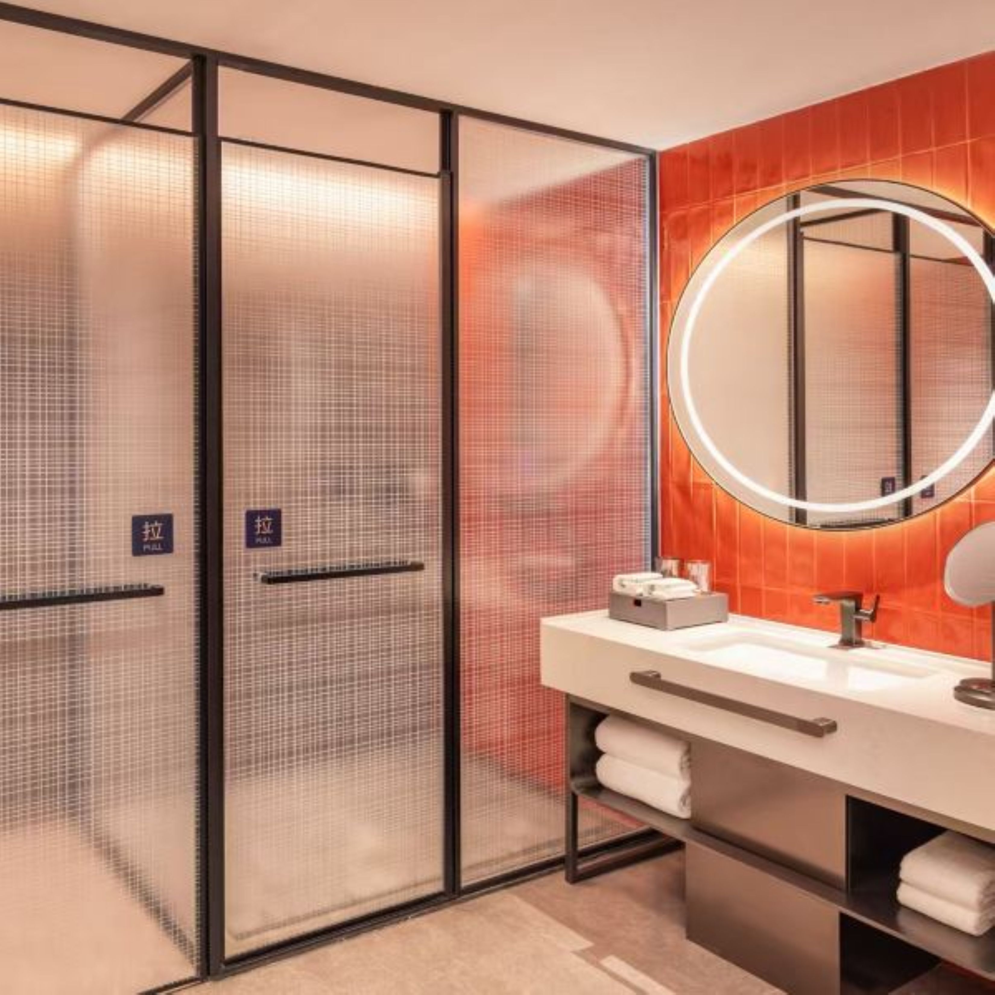 Un bagno moderno con un divisorio in vetro, uno specchio rotondo con illuminazione integrata e un lavandino bianco.