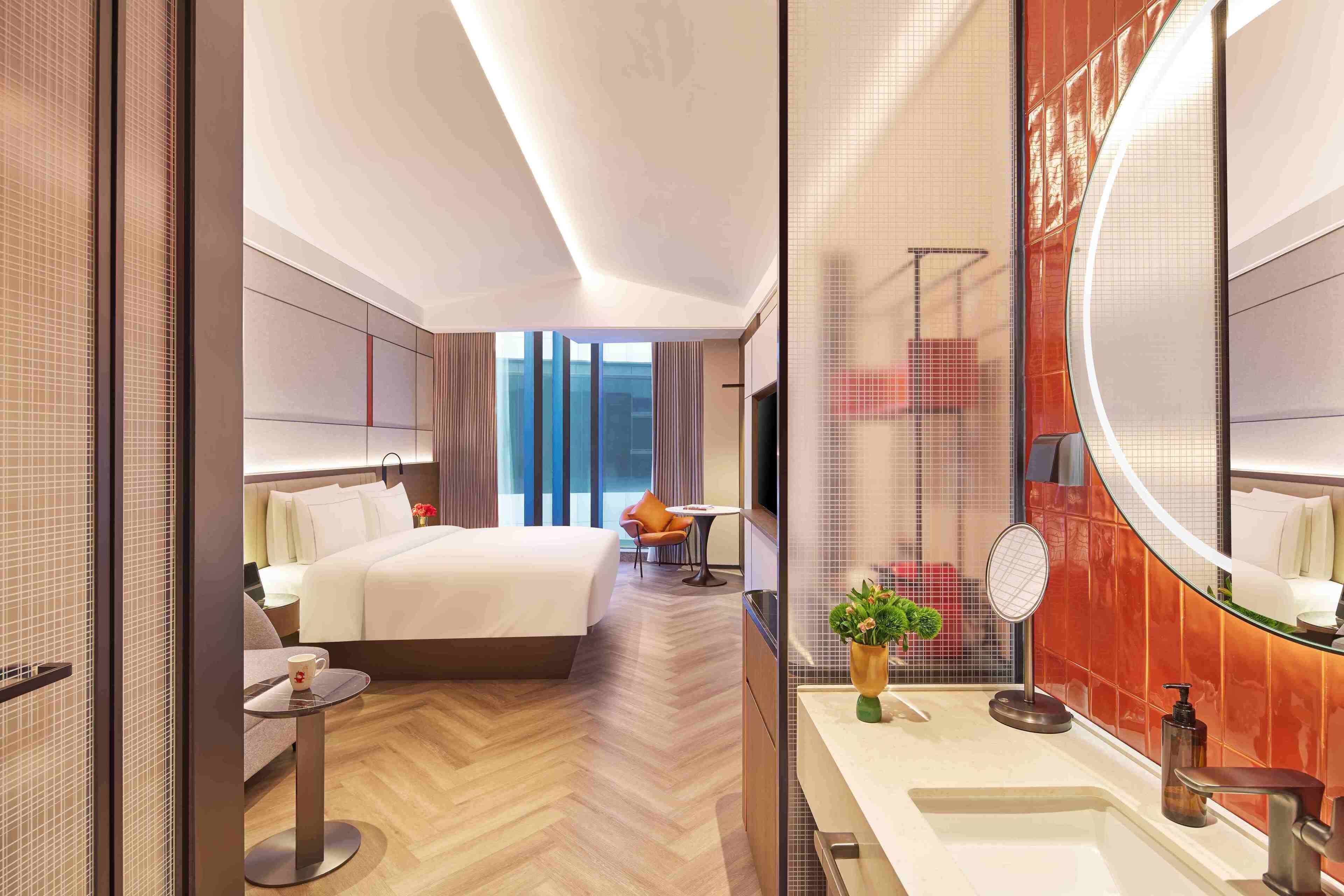 A modern hotel room with a large bed, a sitting area with a table and chair, a bathroom with a large mirror and sink, and a view of the city through large windows.