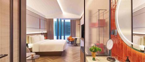 A modern hotel room with a large bed, a sitting area with a table and chair, a bathroom with a large mirror and sink, and a view of the city through large windows.