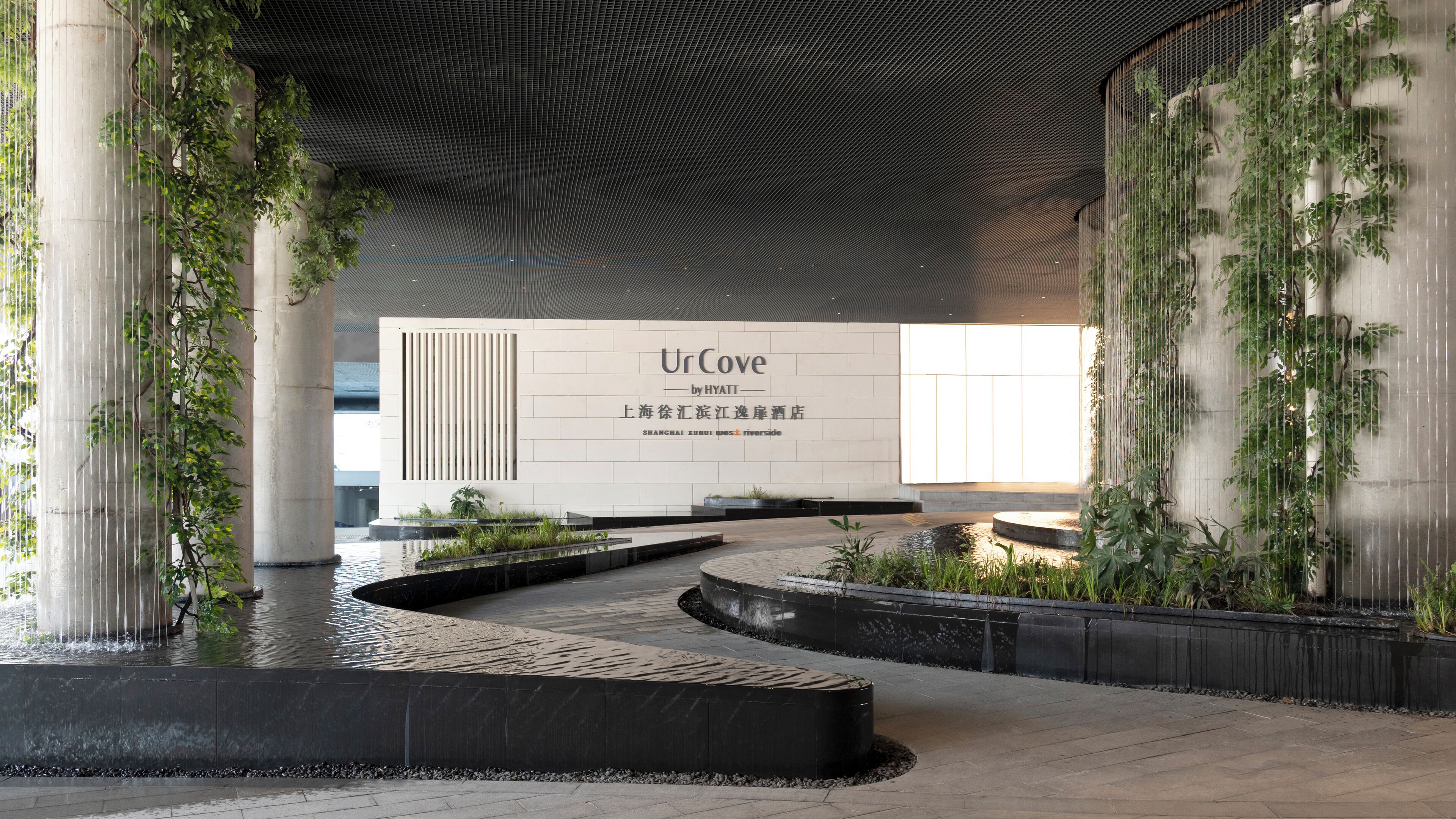 Une entrée d’hôtel moderne, avec une grande enseigne, des éléments décoratifs à base d’eau et de la verdure.
