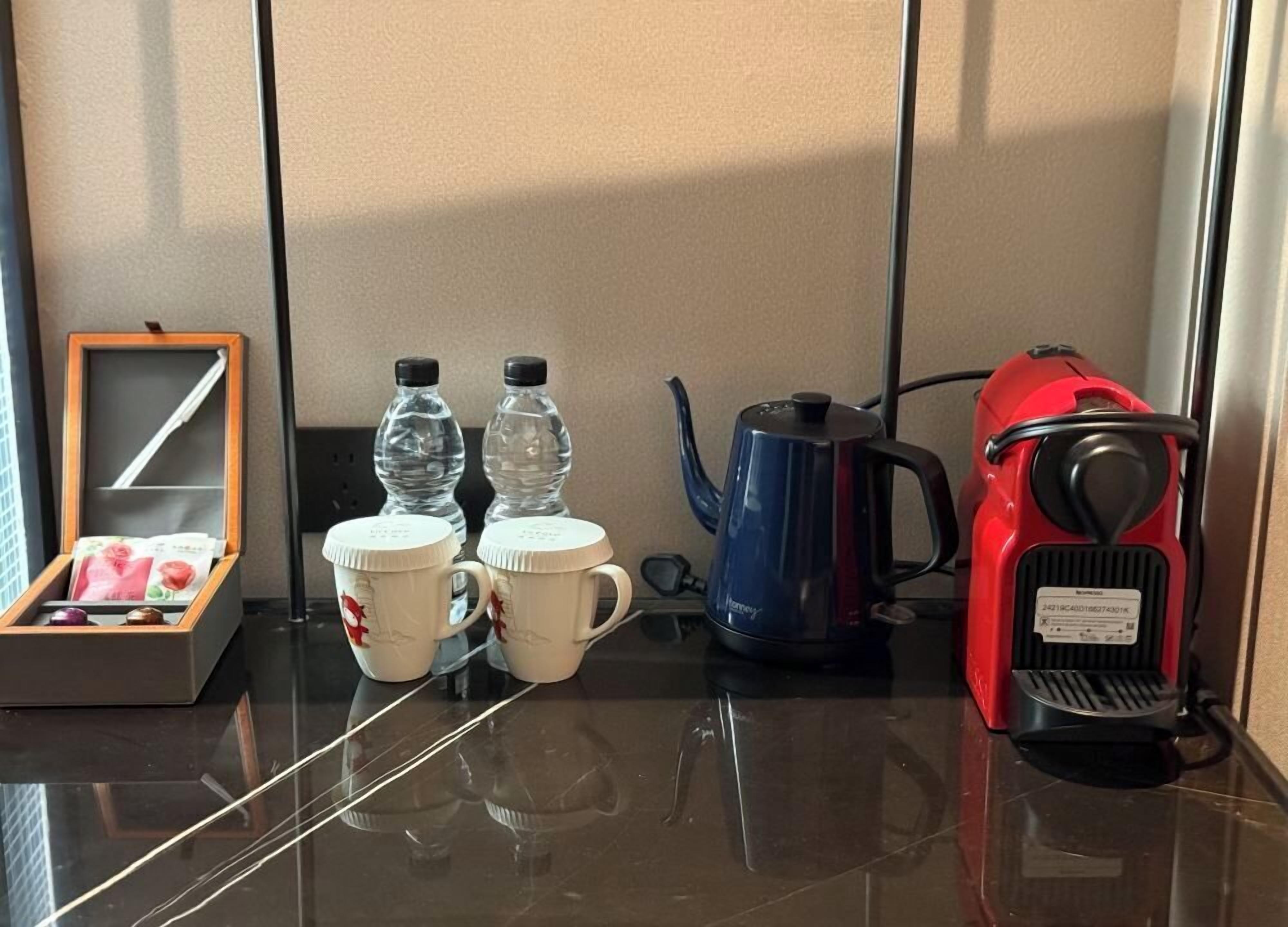 A hotel room with a wooden box, two water bottles, two mugs, a blue kettle, and a red vacuum cleaner on a reflective surface.
