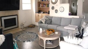 A living room with a grey sofa, a coffee table, a flat-screen TV, a fireplace, a bookshelf, and a kitchen area in the background.