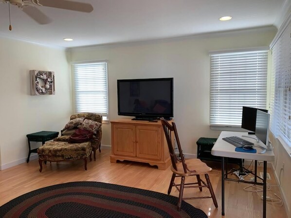 Un salon comprenant un téléviseur à écran plat, un meuble en bois, un fauteuil à motifs, une petite table et un bureau avec un ordinateur.