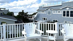 Ein Balkon mit weißen Adirondack-Stühlen und einem kleinen runden Tisch, der auf ein Wohngebiet mit einem grauen Haus und Blick auf das Wasser blickt.