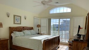 Ein Schlafzimmer mit einem großen Bett, einem Deckenventilator, einem Fenster mit Jalousien, einem Schreibtisch mit Lampe und Blick nach draußen.