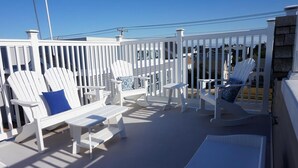 Un espace extérieur aménagé avec des sièges, protégé par un toit, comprenant des chaises Adirondack blanches et une petite table, le tout entouré d’une clôture blanche.