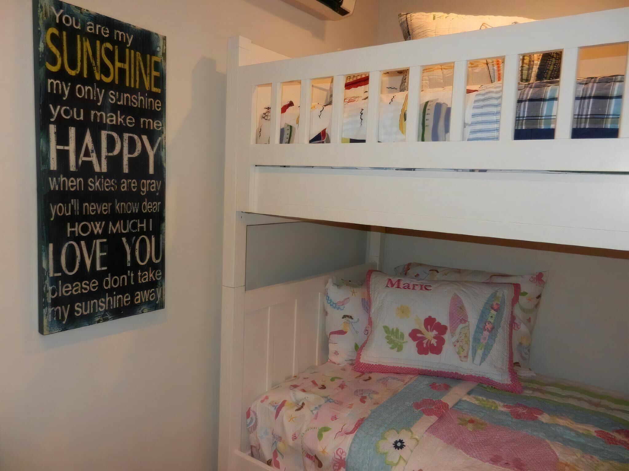 A bunk bed with a lower bunk featuring a floral-patterned pillow labeled 'Marie.' Above the bed, a wall-mounted shelf displays folded clothes. A motivational quote is visible on the wall to the left.