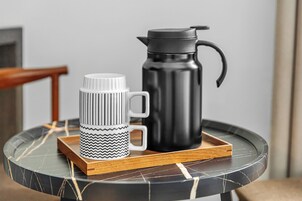 Una taza negra y blanca y un termo negro sobre una bandeja de madera, en una mesa de mármol.
