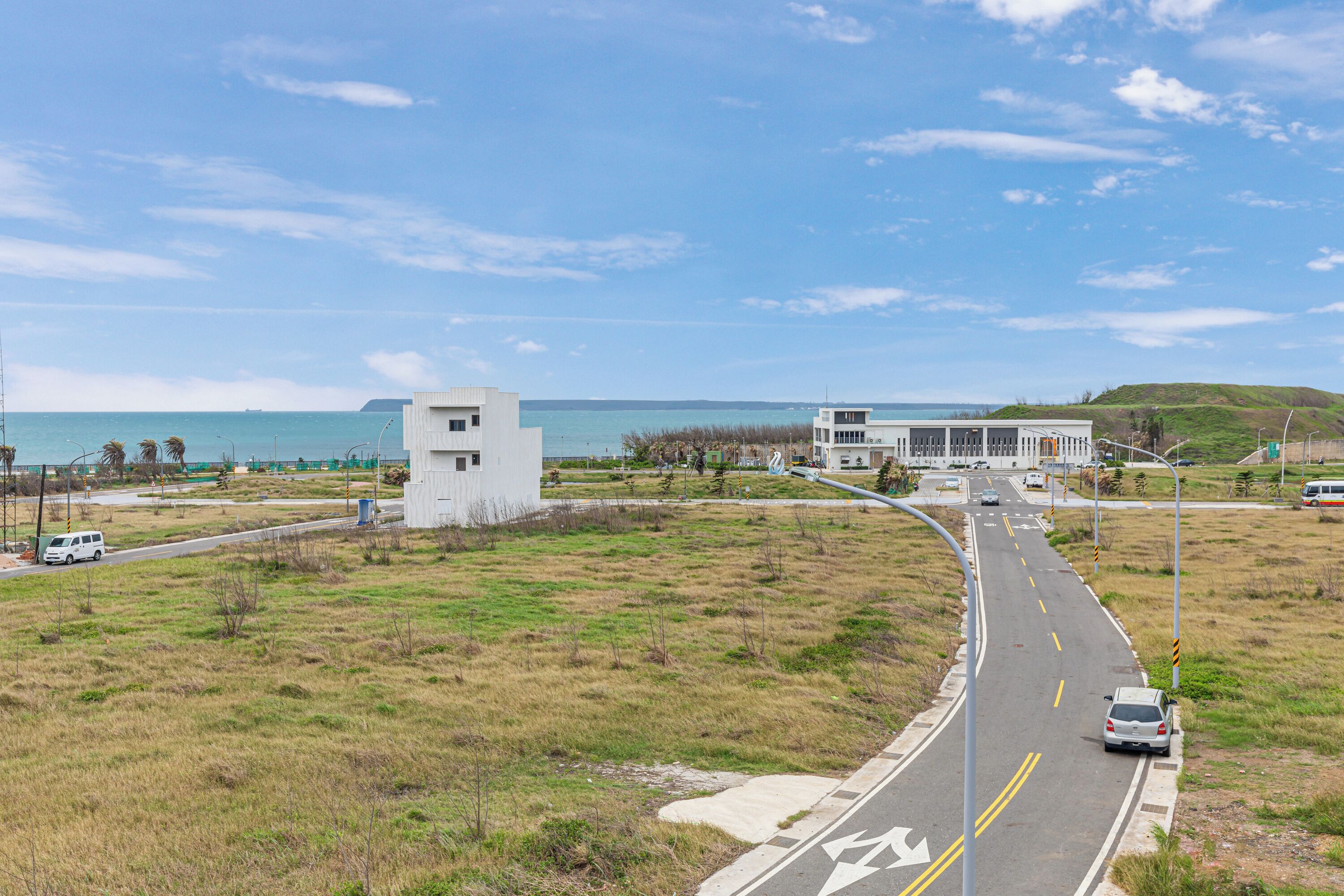 A coastal road with a white building, a grassy area, and a clear view of the ocean.