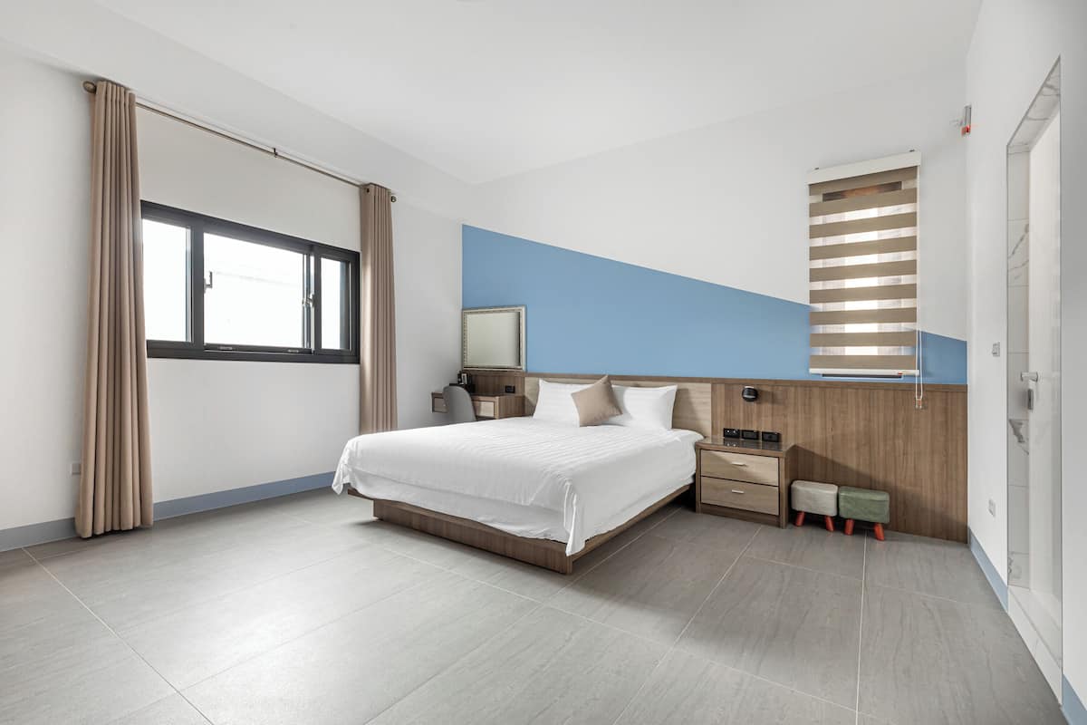 A modern hotel room with a large bed, a nightstand, a wooden headboard, a window with curtains, and a small seating area with a bench and a table.